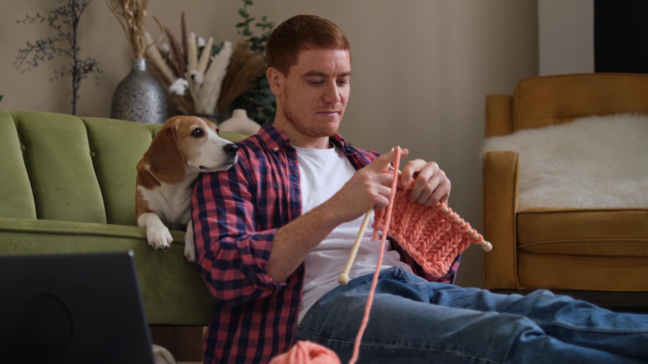 Um homem tricota sentado no chão. Seu beagle está sentado no sofá atrás dele.