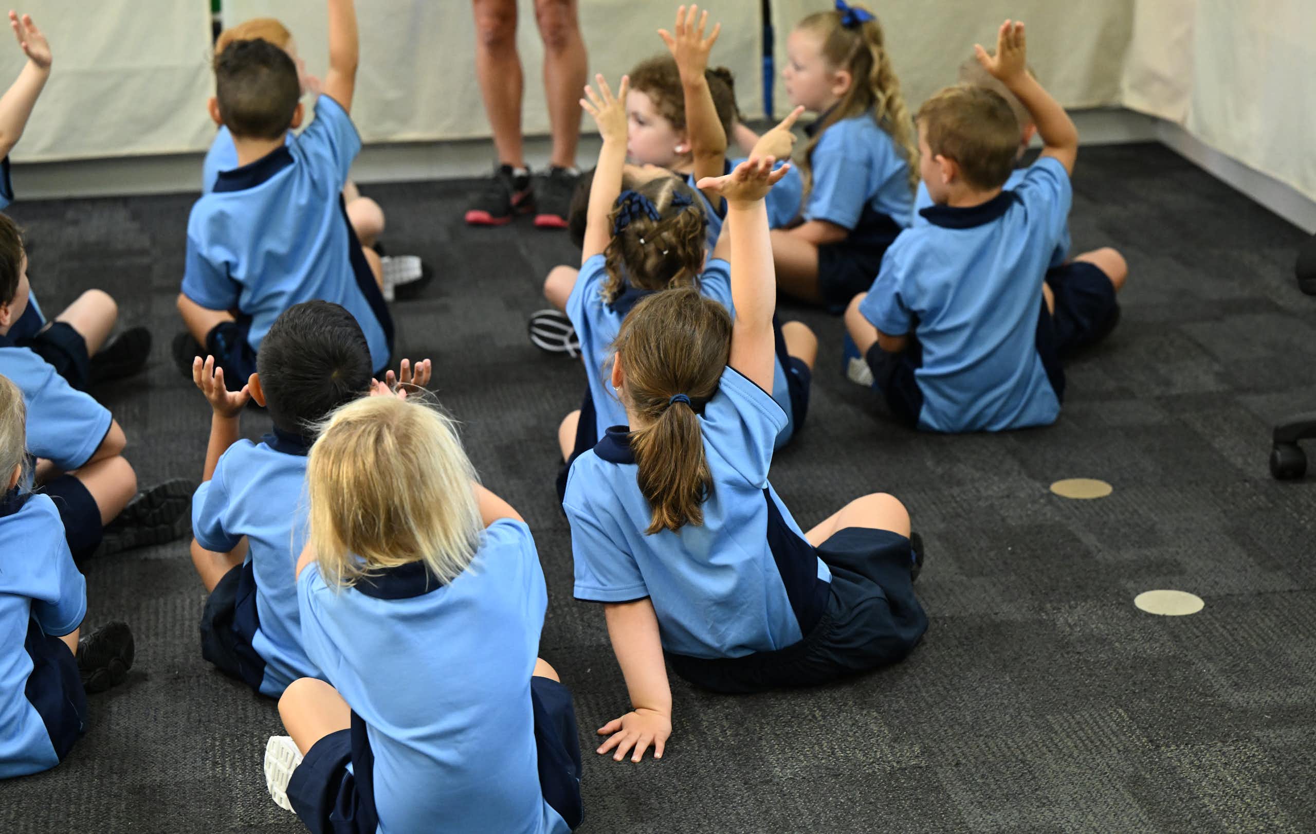 Young primary school children sit on the floor in uniforms. Some have their hands raised. 