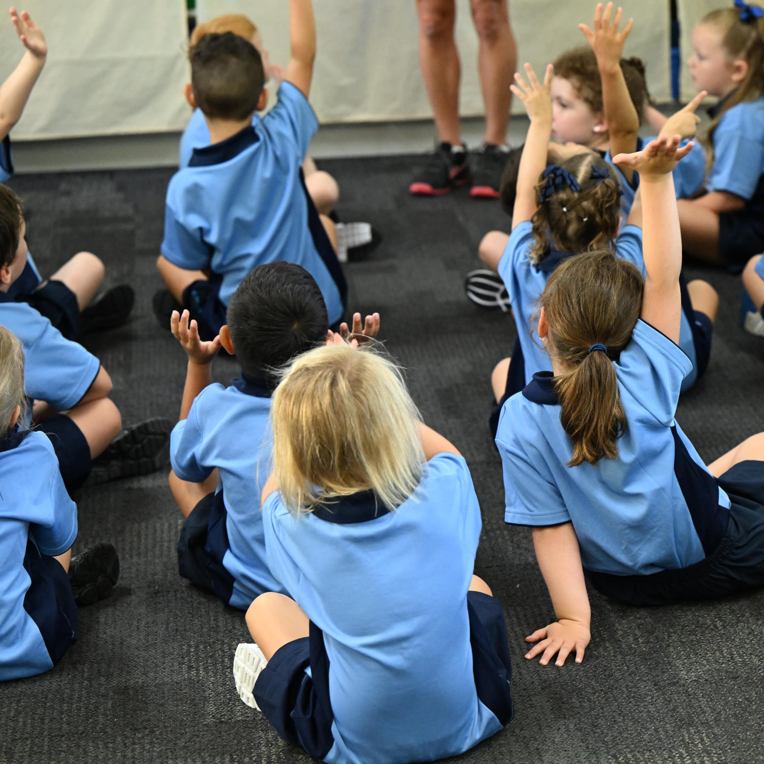 Young primary school children sit on the floor in uniforms. Some have their hands raised. 