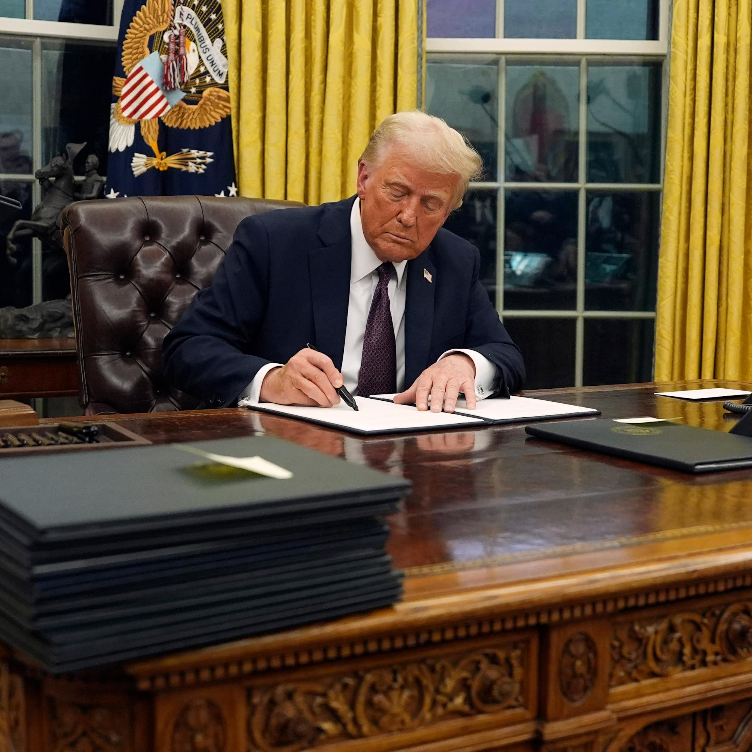 A man sitting at an ornate desk signing a document while another man in a dark suit and red tie stands beside him