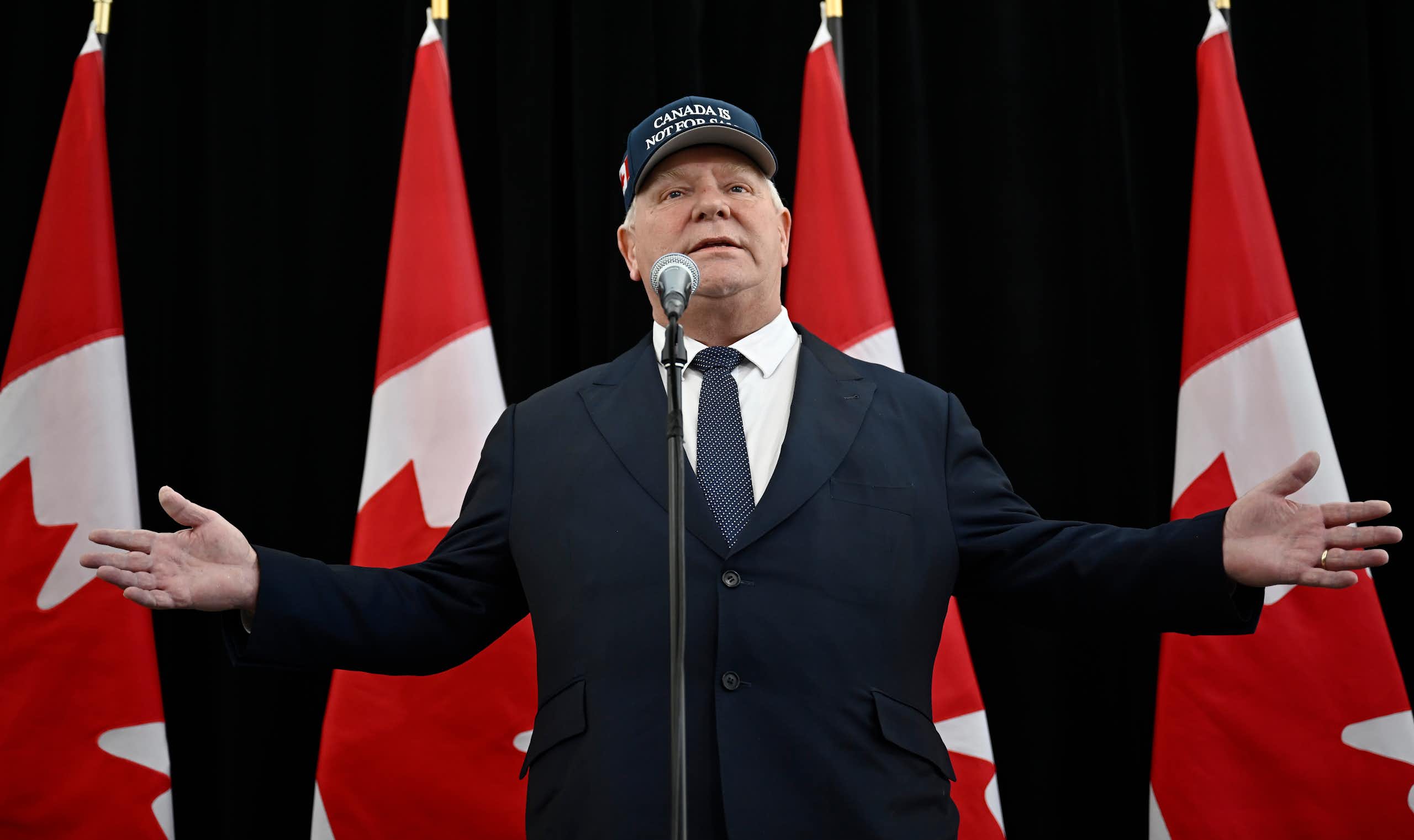 A rotund man stands in front of a microphone with his arms spread wearing a Canada is Not For Sale hat.  A row of Canadian flags are behind him.