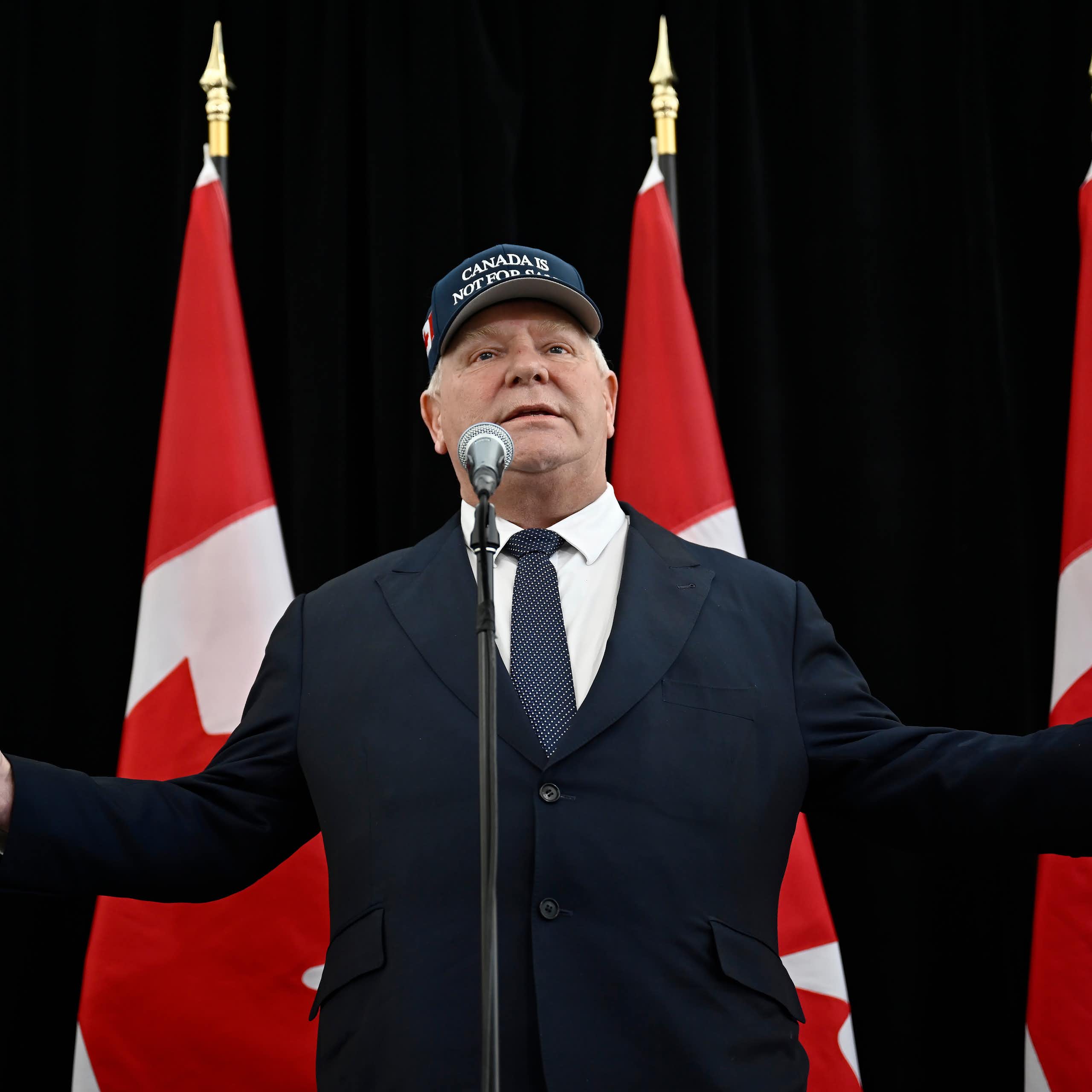 A rotund man stands in front of a microphone with his arms spread wearing a Canada is Not For Sale hat.  A row of Canadian flags are behind him.