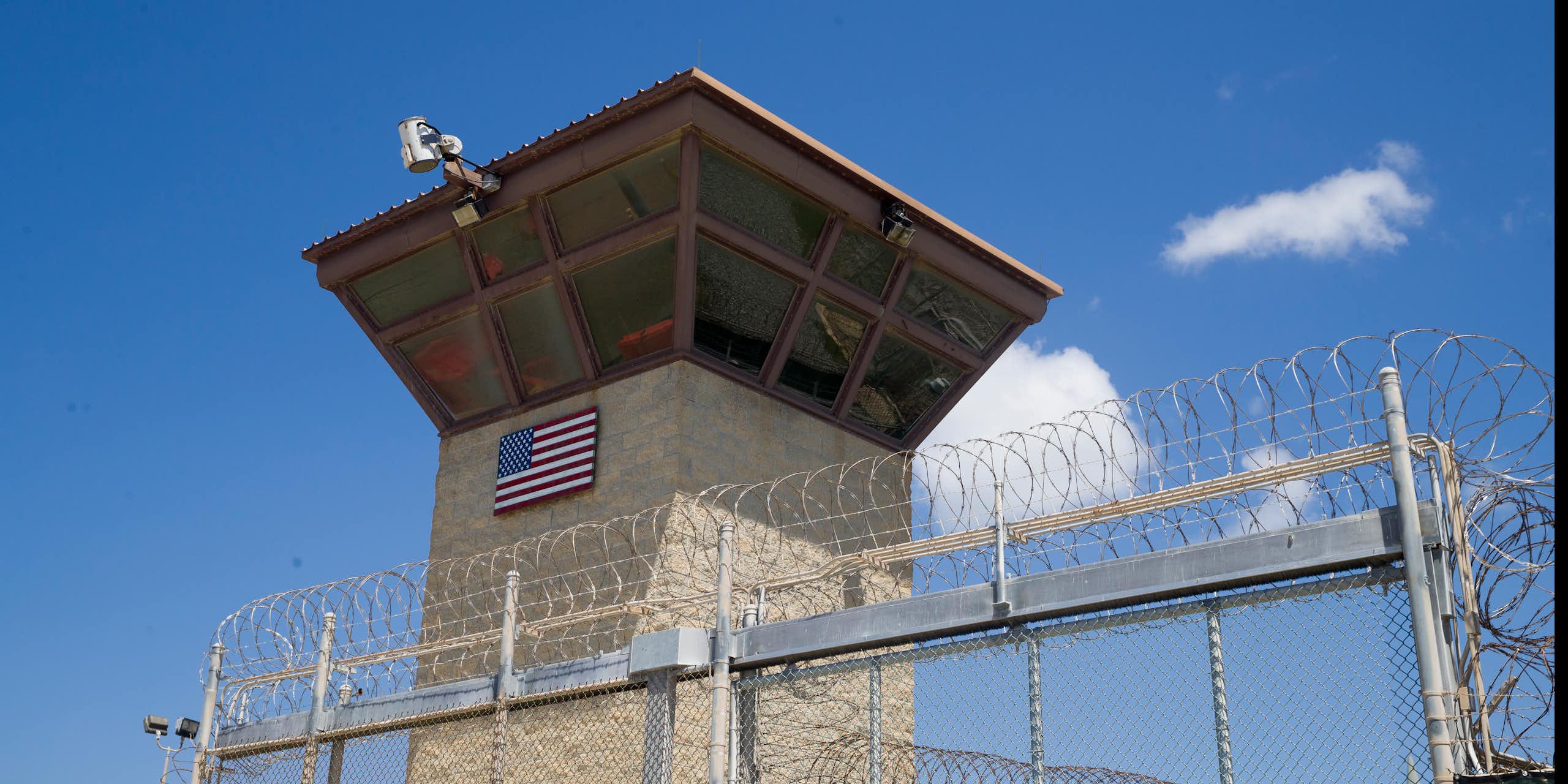 A large tower with windows and a camera stands behind a tall fence topped with barbed wire.