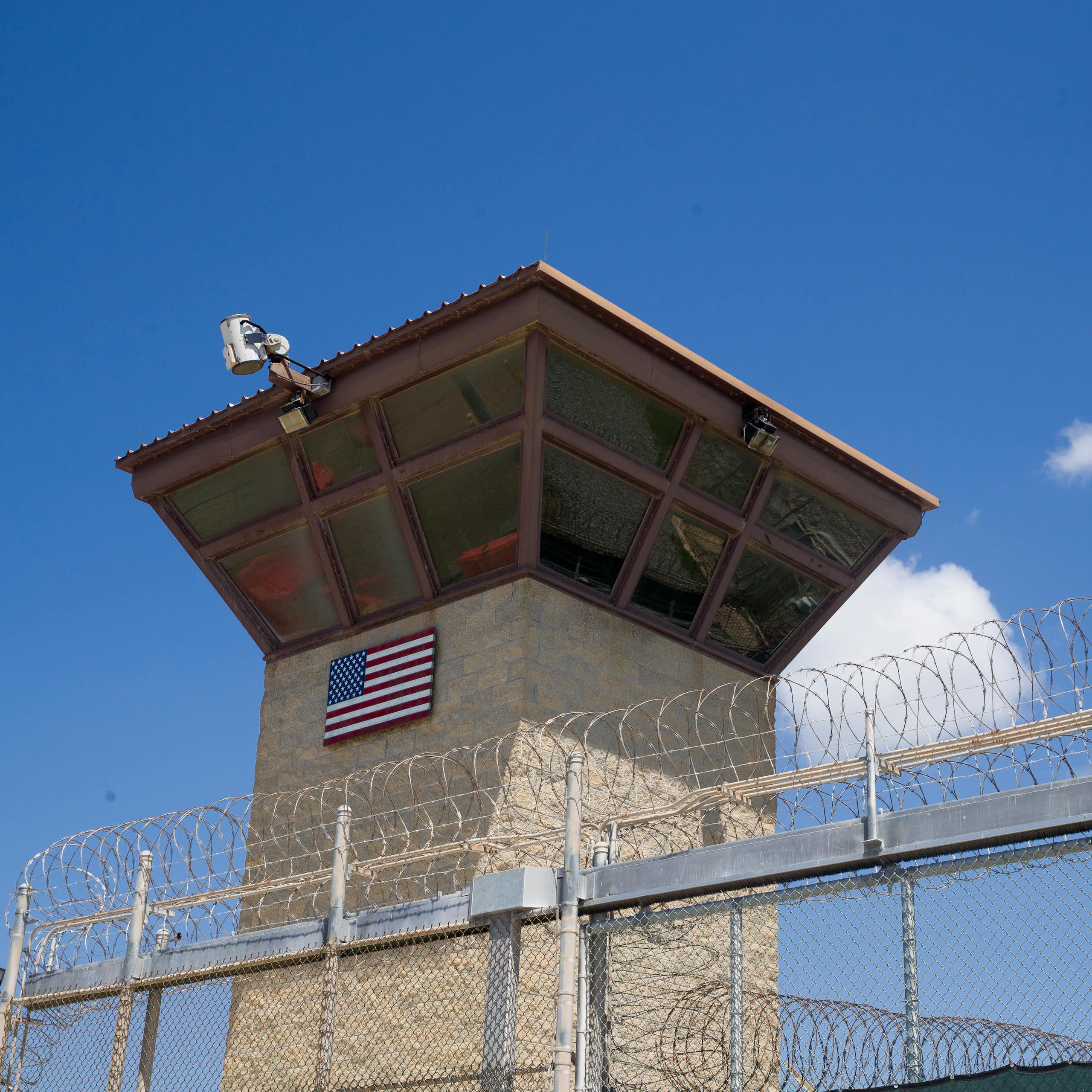 A large tower with windows and a camera stands behind a tall fence topped with barbed wire.