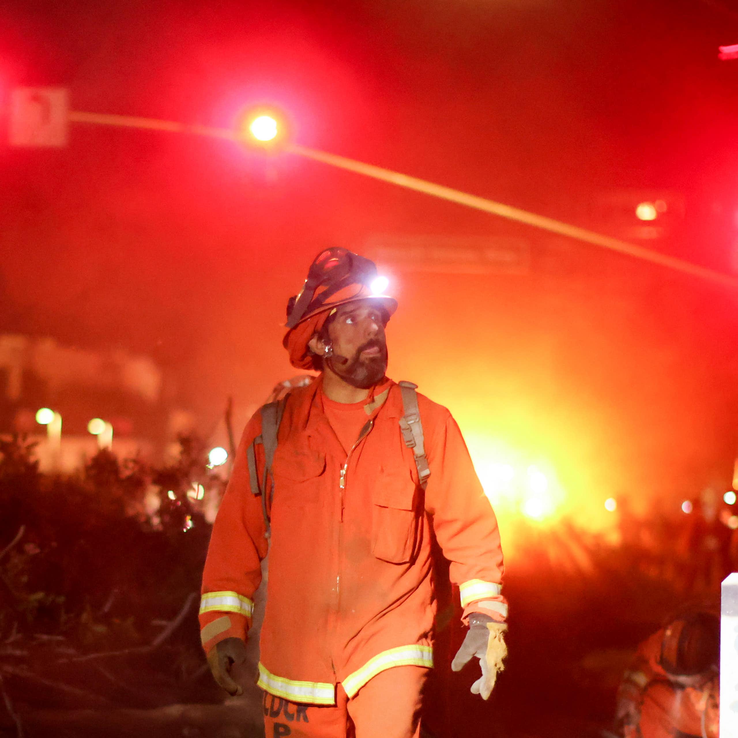 A man in an orange firefighting uniform and hard hat walks down a street. An orange glow is seen behind him