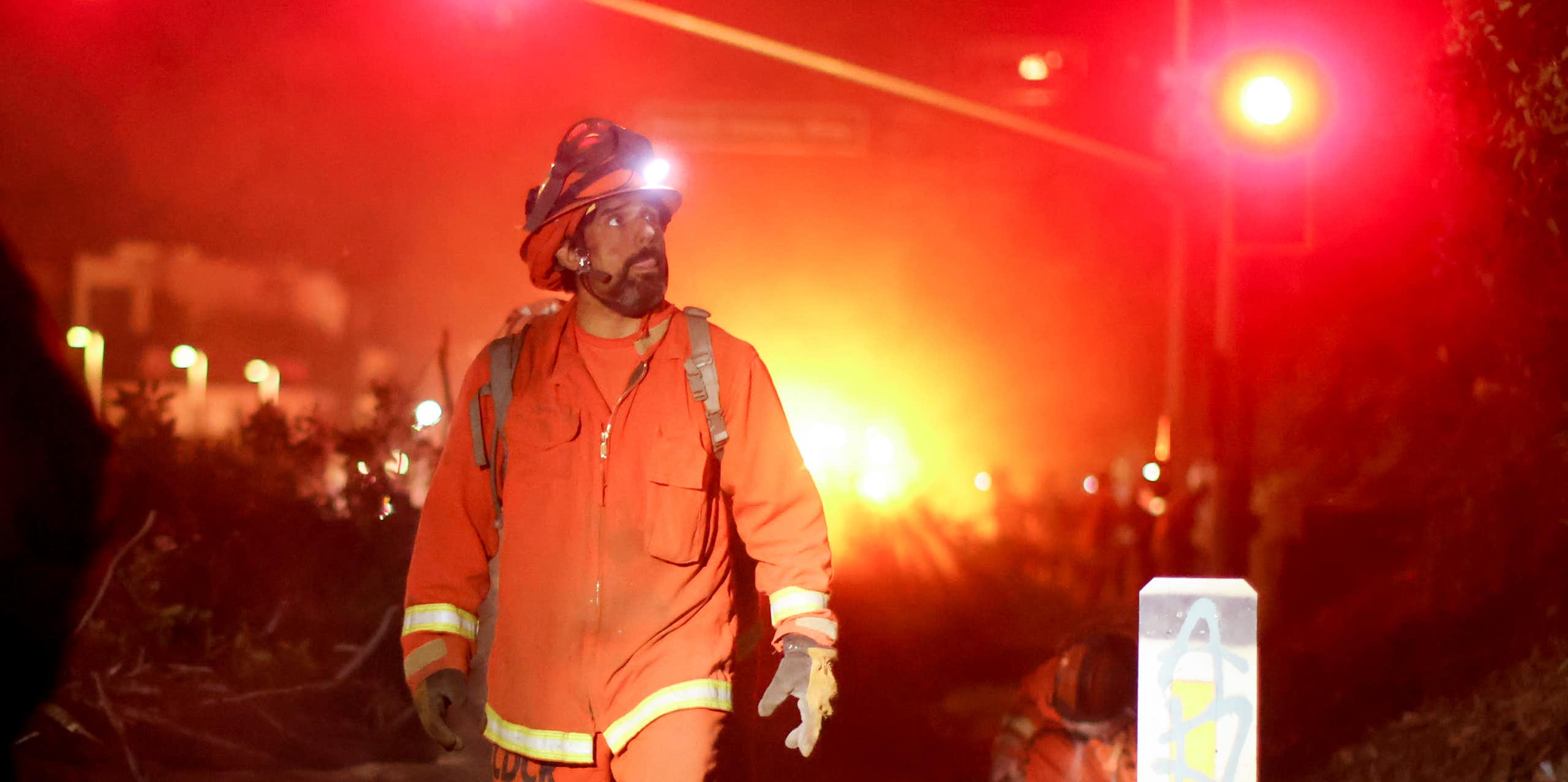 A man in an orange firefighting uniform and hard hat walks down a street. An orange glow is seen behind him