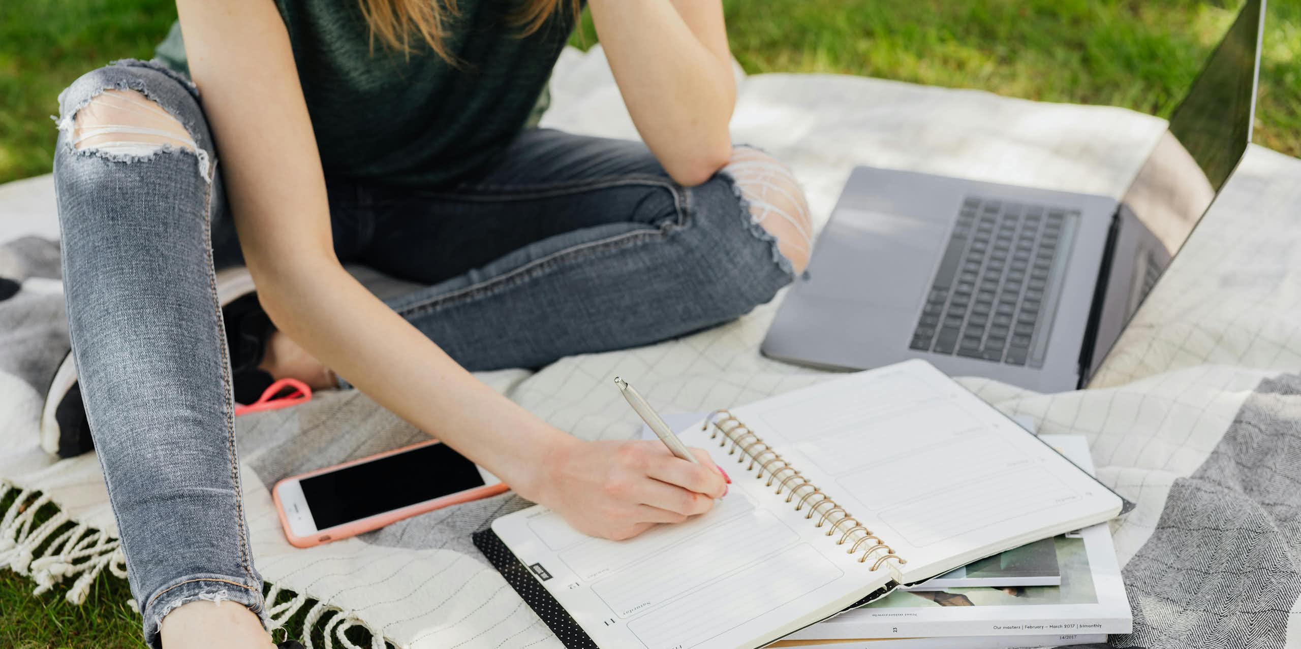 A young person sits on a picnic blanket on grass, with a laptop, planner and notebooks. 