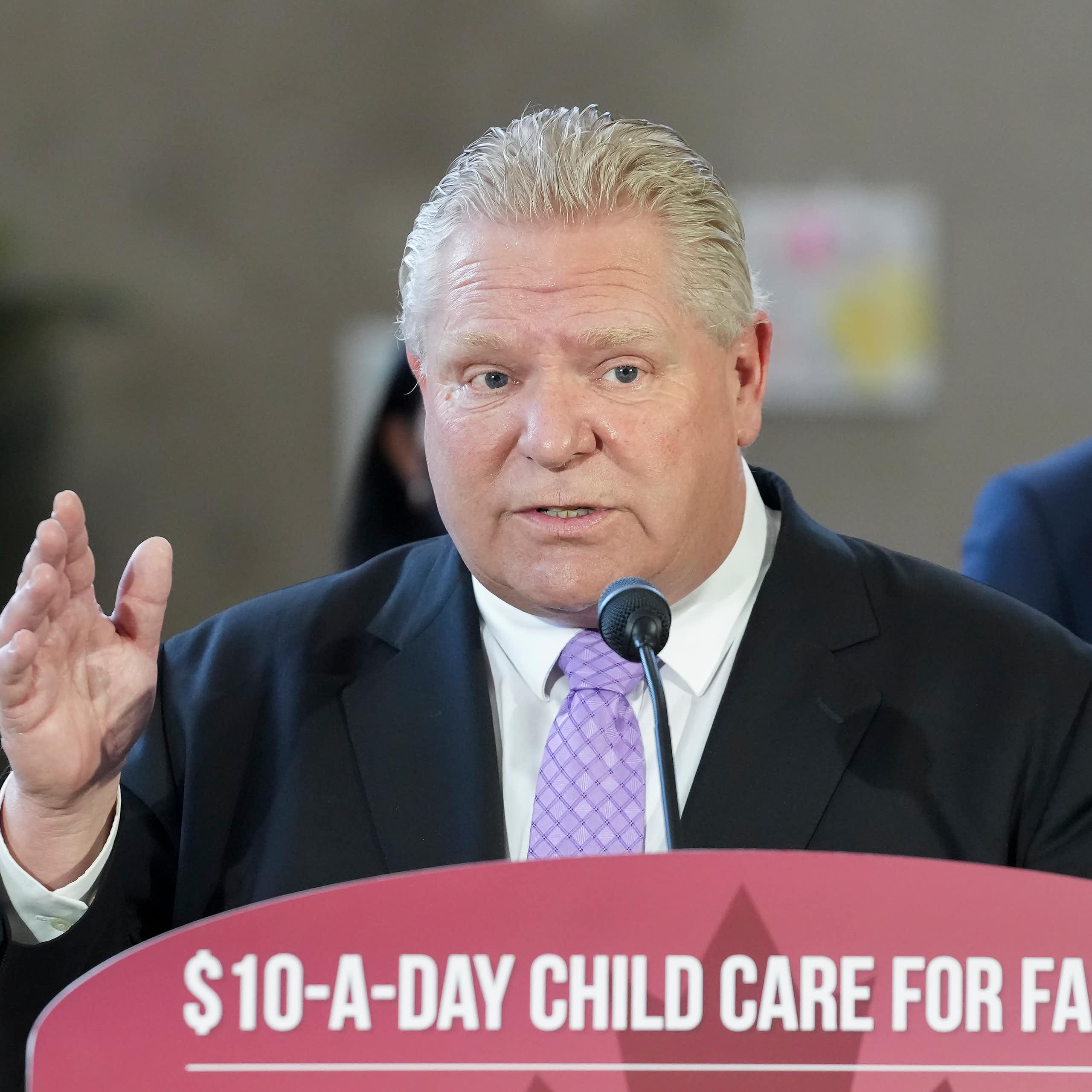 A man at a podium in a suit speaking with a $10 a day child care for families sign.
